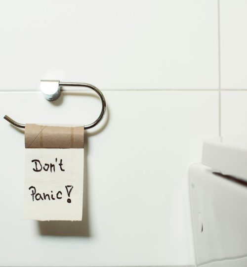 A toilet paper holder with an empty roll displaying a 'Don't Panic!' note, set in a modern bathroom.